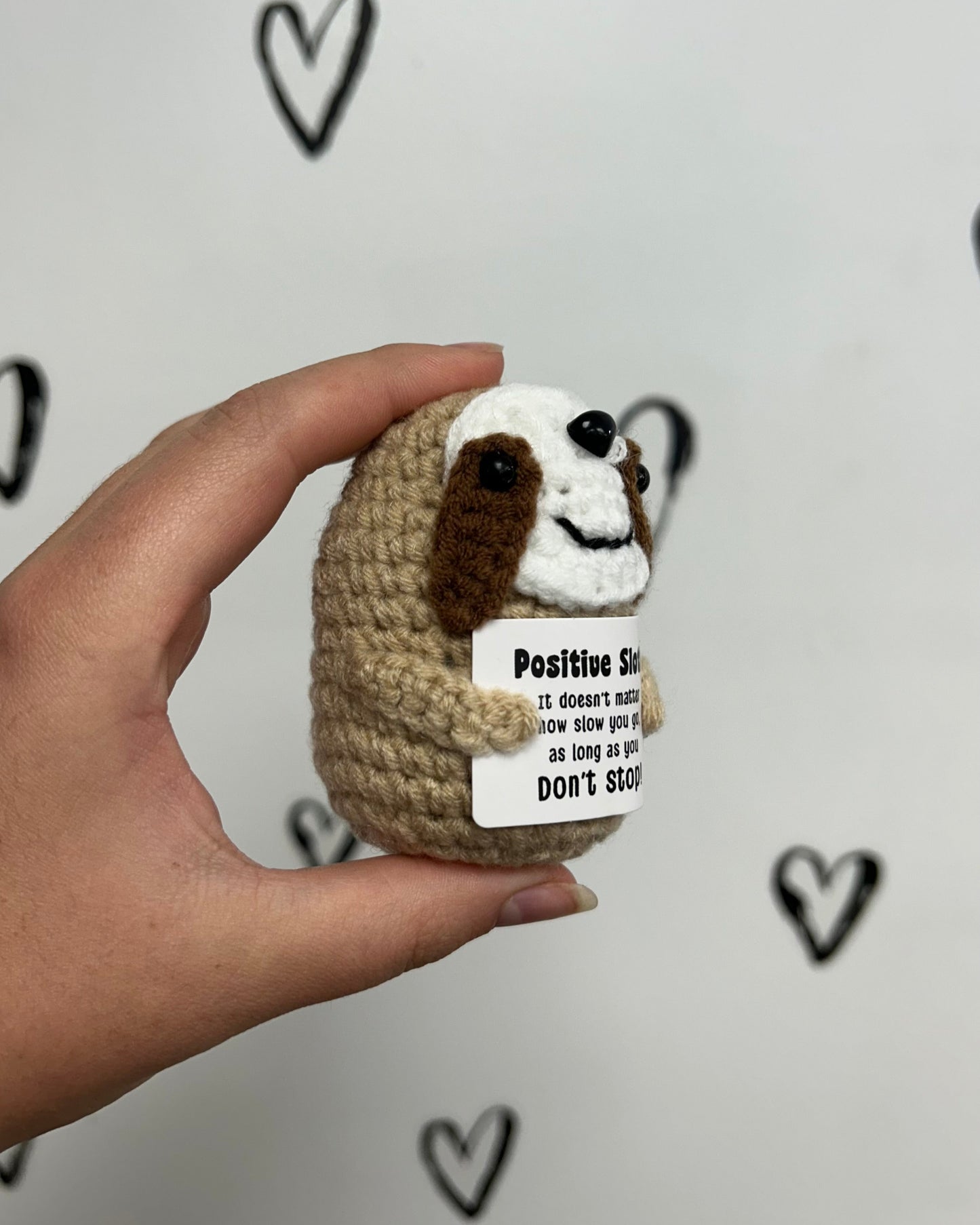 Hand holding a small crocheted animal figure with a motivational tag against a light background with heart shapes.