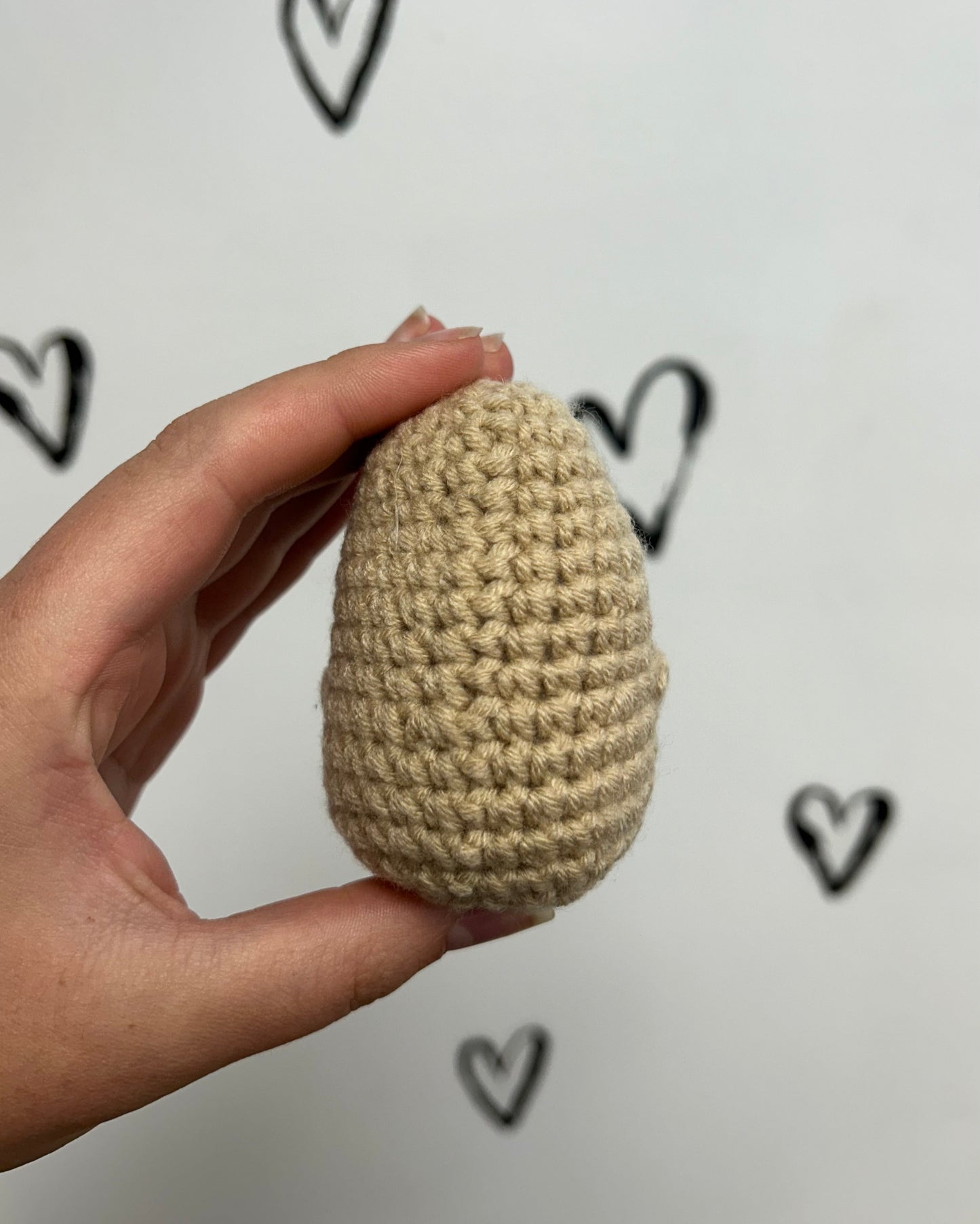 Hand holding a beige crocheted egg against a white background with black heart shapes.