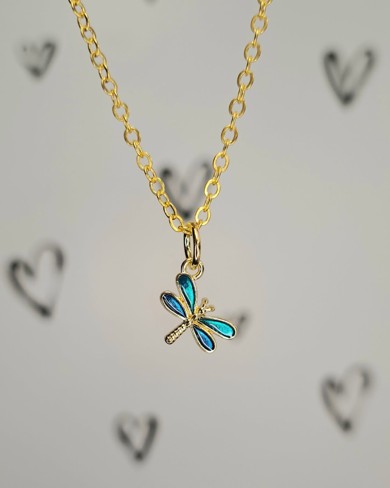 Gold necklace with a blue dragonfly pendant on a light gray background with heart shapes.