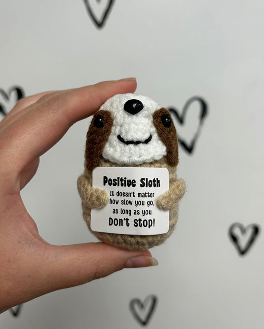 Hand holding a small crochet sloth toy with a motivational message card against a white background with heart shapes.